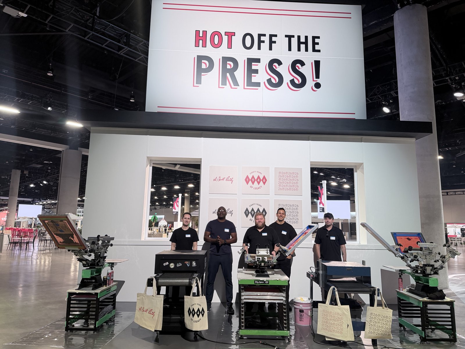 Merch Troop crew standing behind a row of heat presses with pressed canvas totes displayed in front